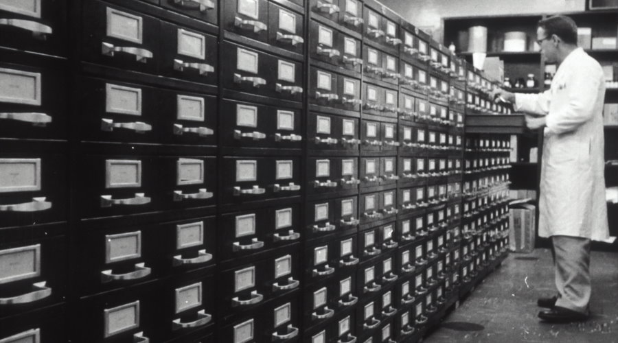 Man in white lab coat searches through records in an archive (black and white)