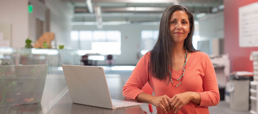 Professional woman in an orange top leans against a desk next to a laptop